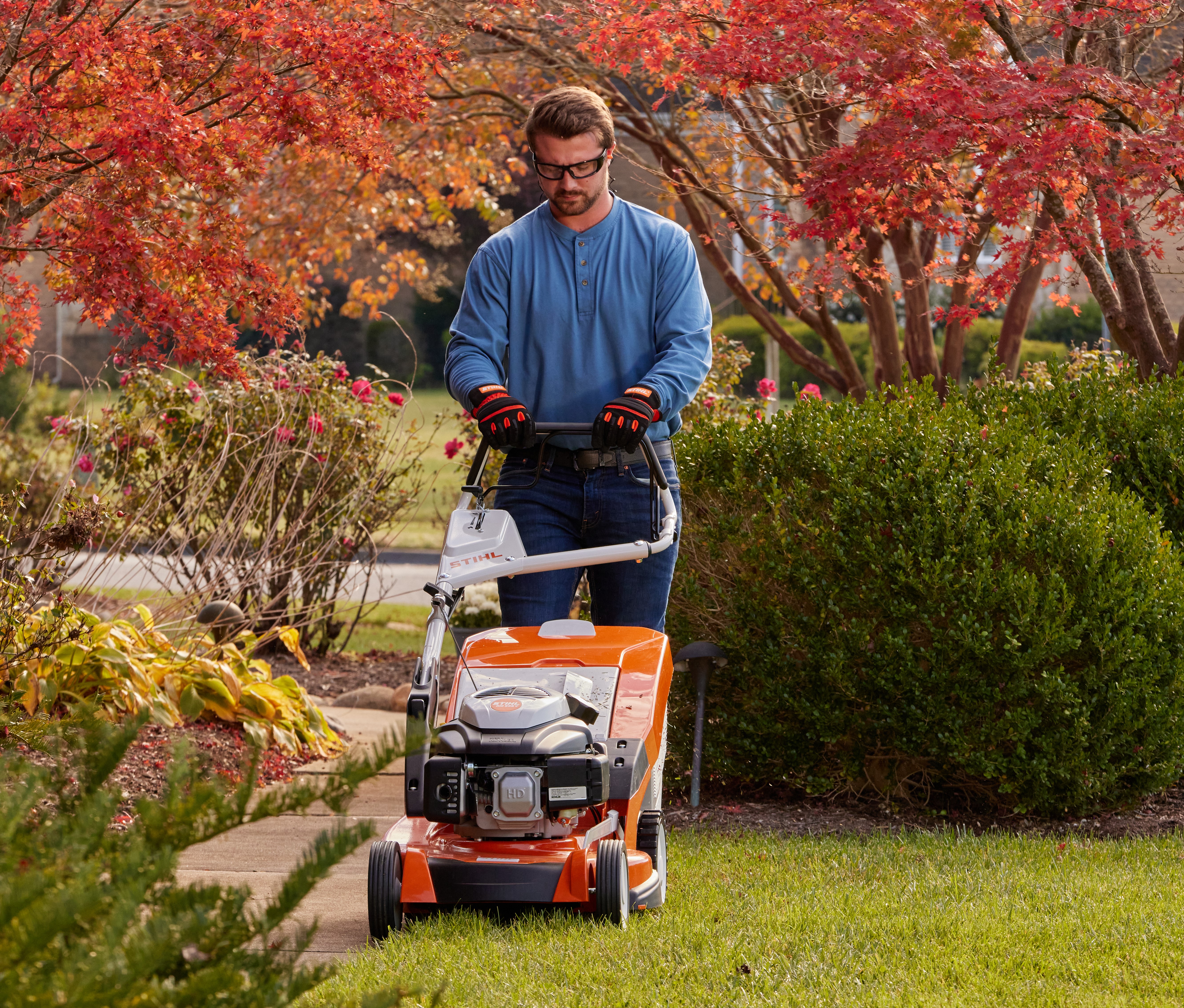 A man is halfway through mowing untidy and overgrown grass using a STIHL RMA 448 P-C cordless lawn mower, in a garden surrounded by tall shrubs and plants
