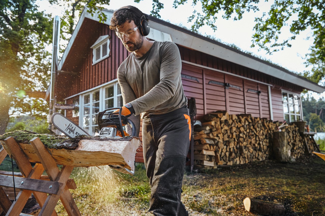 Man sitting on a sawhorse in front of a pile of split logs.