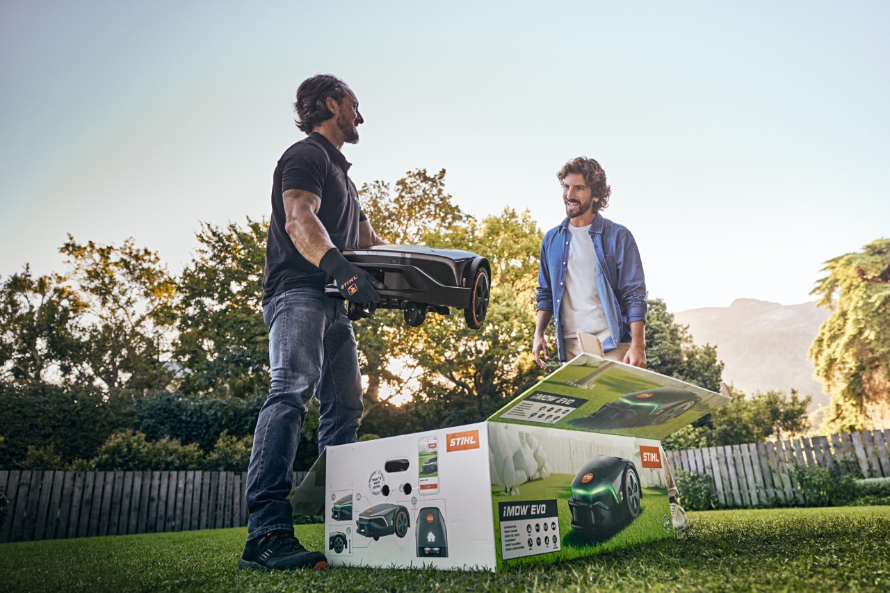 A STIHL dealer standing in a customer’s garden with an iMOW® EVO in their hand and the iMOW® box in front of them, ready to help with installation