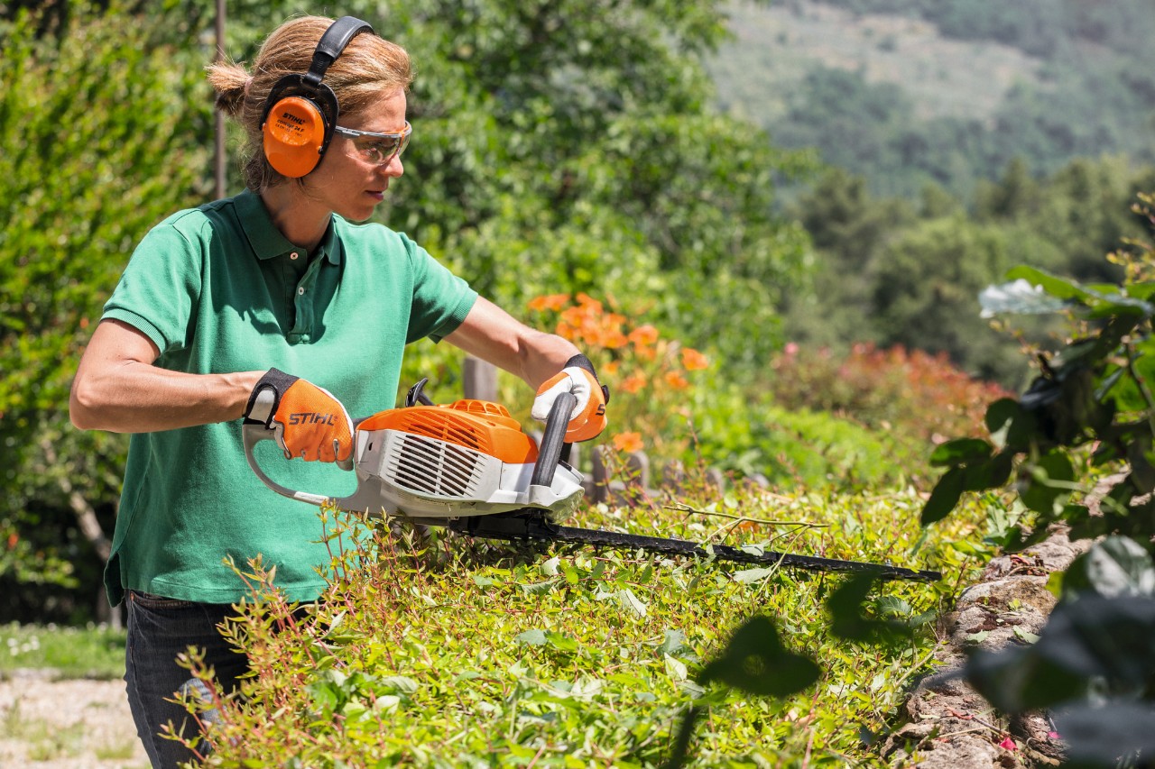 Femme portant des protections auditives et des lunettes de protection coupant une haie avec un taille-haie à essence STIHL HS 46