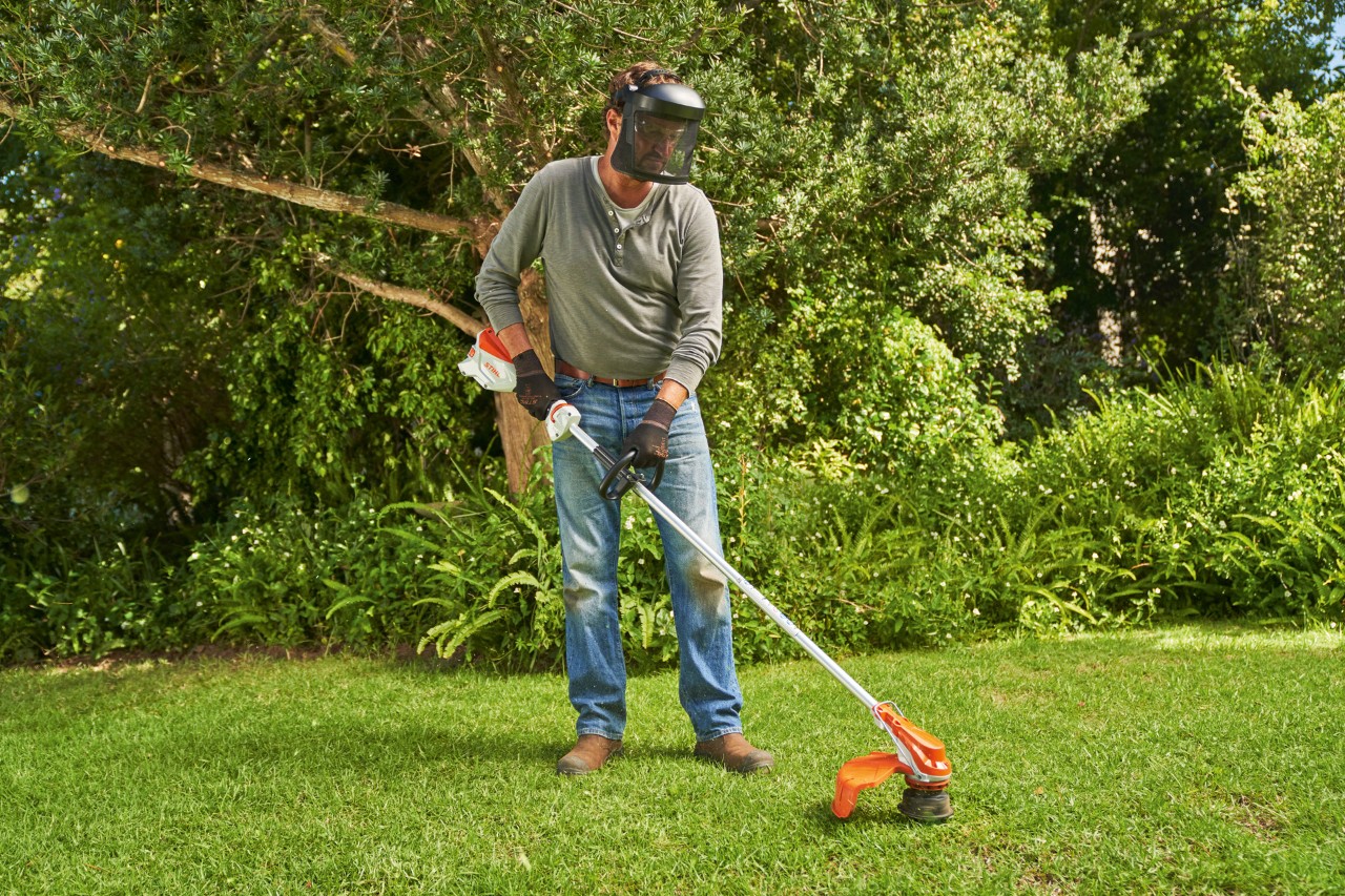 Un homme portant une visière et tondant la pelouse dans un jardin à l’aide de la débroussailleuse à batterie STIHL&nbsp;FSA&nbsp;60